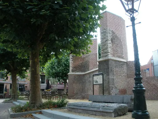 The image shows a historical structure, likely a ruin or part of a building, made of brick. Surrounding the structure are trees and a lamp post, suggesting it is in a garden or public space. There is also a sign nearby that may provide information about the site. Additionally, there seems to be outdoor seating or tables in the background, indicating it might be near a café or restaurant.

Image Credits: Wikimedia / Jvhertum / Public domain
