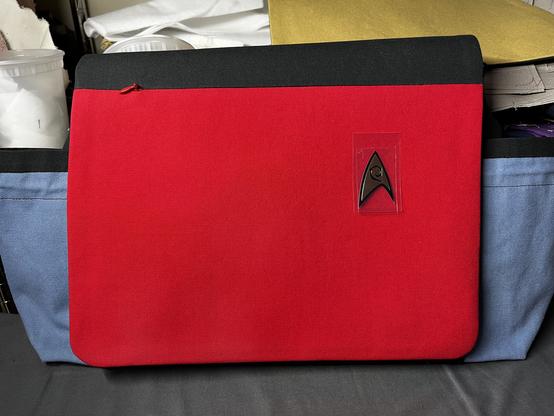 A red messenger bag flap with a black upper band is draped over a powder blue bag where the side pockets are visible. There is a small (~2”) badge in the top left corner, positioned as it would be on a Starfleet uniform from TOS.