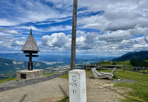Dreiländereck mit dem Grenzstein mittig, einem Wegmarterl und Sitzbänken mit Blick auf Villach und dem Wörthersee