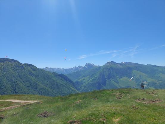 Le pic soum de Pirait en face, depuis la zone de décollage parapente. Ciel dégagé grand soleil petite brise du nord / nord ouest qui s'installe visible grâce à la manche à air. Multiples ailes colorées regroupées dans la masse d'air cherchant les instabilités aériennes pour monter