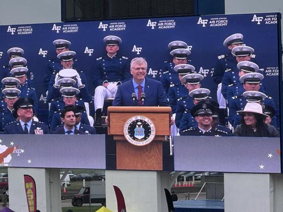 An outdoor ceremony featuring a speaker at a podium in front of a large screen displaying the United States Air Force Academy logo. In the background, a group of uniformed Air Force personnel, including cadets and officers, is seated.