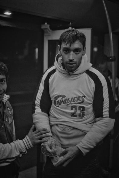 The black and white portrait-orientated image shows a man in a hoodie holding a plastic bag in his hands, which are clasped together in front of him. He is looking directly towards the camera. The man's hoodie is light-colored with dark sleeves and features the word "CAVALIERS" and the number "23" on the front, along with some other text below it. To the left of the man, another individual's arm and a partial view of their body are visible. The arm is reaching towards the man's hands. The scene is on public transportation, with a dark background and some faint reflections or lights visible.