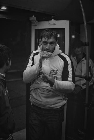 The black and white portrait-orientated photo features a man in a hoodie, holding a plastic bag up to his mouth. The bag is half full with light-colored content. His eyes are looking towards the camera. The hoodie is light-colored with dark sleeves and bears the text "ERS". Another individual's back is partially visible on the left side of the frame, facing away from the camera. To the right and behind the man, another person is faintly visible. The background is dark, the scene is on public transportation, with a doorway behind the man.