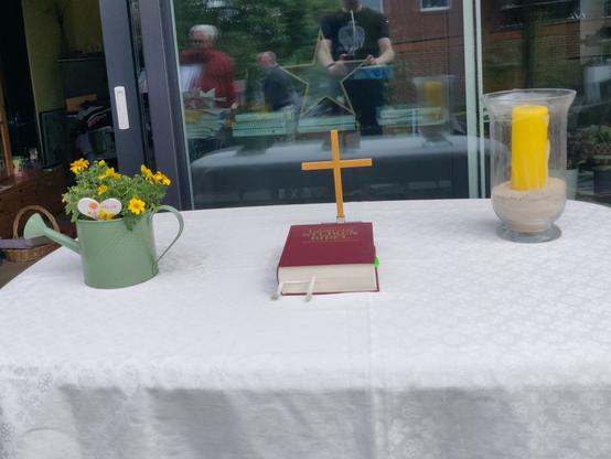 Ein improvisierter Abendmahlstisch, weiße Decke, Bibel, Kreuz, Blumen und eine Kerze, vor einem großen reflektierenden Fenster.