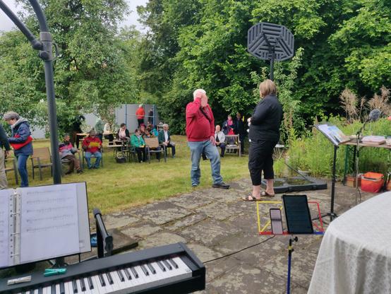 Eine Sandstein-Terrasse, Rasen mit Bäumen, darauf Bänke mit mehreren Besuchern, zwei Personen unterhalten sich im Vordergrund. Links vorne ein Digitalpiano, rechts ein Tisch, ein Notenpult und ein Sprecher-Mikrofon mit Windschutz.
