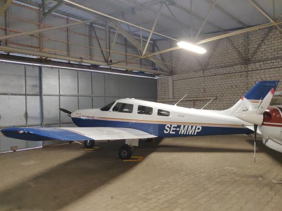 A white-and-blue low-wing light aircraft sitting in a hangar under fluorescent light.
