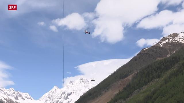 Aufnahme des Schweizer Fernsehen SRF eines Autos, das an einem Hubschrauber hängt, inmitten einer hochalpinen Gebirgslandschaft. Am unteren Bildrand sind massive Bergrutschungen zu erkennen.