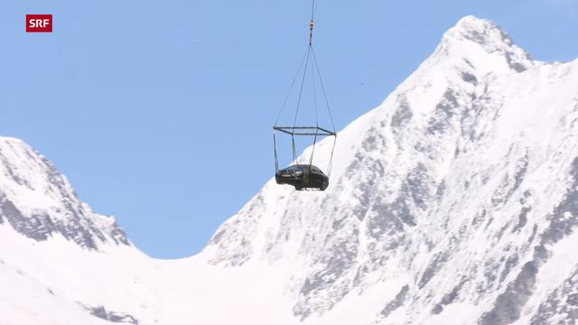 Aufnahme des Schweizer Fernsehen SRF eines Autos, das an einem Hubschrauber hängt, inmitten einer hochalpinen Gebirgslandschaft mit verschneiten Gifpeln.