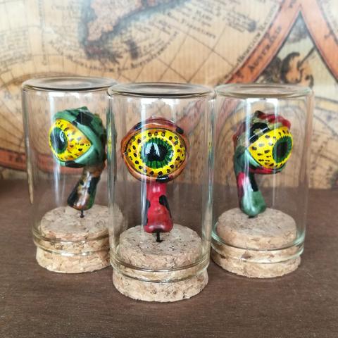 Un œil au bout d'un pédoncule dans une petite fiole en verre de 30mm de diamètre. Il est jaune avec des tache autour de l'iris comme ceux de certains batraciens.