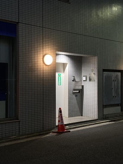 An illuminated entrance to a building at night, featuring a circular light and a green emergency exit sign. There is a traffic cone positioned near the entrance, and the wall is tiled. A mailbox is visible next to the entrance.