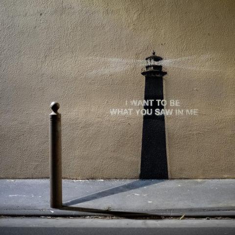 Stencil artwork by Banksy on a beige wall resembling a lighthouse, with a cleverly integrated bollard on the sidewalk completing the base of the tower. The light beam from the top of the lighthouse cuts through the text: “I want to be what you saw in me.”