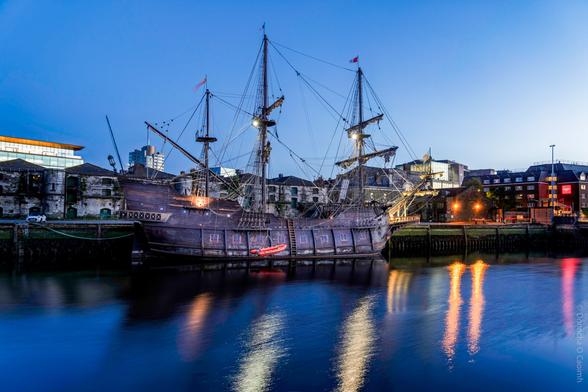 A full-scale replica of a 16th–17th century Spanish galleon, the Galeón Andalucía, docked at dusk along the quay in Cork, Ireland, with its tall masts, intricate rigging, and wooden hull reflected in the calm river water; city buildings and soft evening lights form the backdrop.