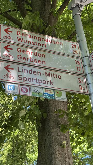 Wegweiser mit 6 Zielen an Pfahl vor Laubbaum bei Sonnenschein. Oben: Barsinghausen 29 km