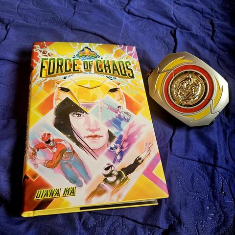 Buch 'Force of Chaos' über Mighty Morphin Power Rangers von Diana Ma neben dem Power-Morpher der gelben Rangerin auf blauem Stoff