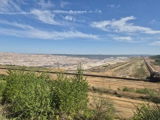 Das Bild zeigt den Tagebau Hambach, ein gewaltiger Braunkohletagebau unter einem klaren, blauen Himmel mit einigen weißen Wolken. Im Vordergrund sind grüne Büsche und Sträucher zu sehen, die einen Kontrast zu der trockenen, erodierten Landschaft bilden. In der Mitte des Bildes erstreckt sich eine riesige, offene Grube. Der Tagebau wird von einem langen, breiten Förderband durchquert, das sich von rechts nach links erstreckt. Im Hintergrund sind Windräder und Kohlekraftwerke zu sehen.