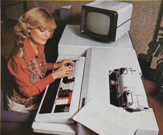 A photo of a woman sitting at a white desk-sized computer, her hands on the keyboard. The entire console appears to be built from two parts: a substantial keyboard and printer unit, and a smaller but still desk-height unit, which is probably the computer itself, concatenated onto the left of the main unit.

In the main unit, a sprocket-fed printer mechanism is built into the desk behind the keyboard. On the smaller unit, a CRT monitor with a black bezel (apparently switched off) is diagonally to the left of the woman. Her attention is on a piece of paper on the desk diagonally to her right. She is smiling serenely.

Her hair is blonde and styled in a medium-length 1970s style, cow's licks aplenty. She is wearing a long-sleeved red blouse and a floral waistcoat in creams, browns and oranges, and white trousers.