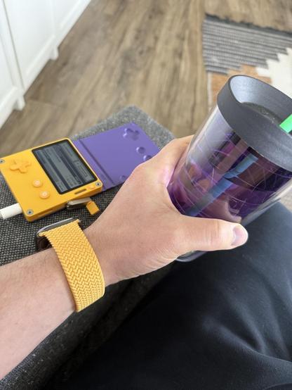 Playdate handheld gaming console in yellow with purple cover on chair arm, next to arm wearing watch with matching yellow wristband, hand holding plastic water cup in matching purple.