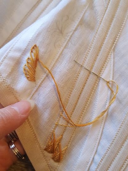 A close up photo of the side front of a white corset with pale tan boning channels, and some embroidery on gold thread being sewn on it. The thread has a slight variegation between pale gold and rose gold, and it is currently becoming a leaf via blanket stitch. There's so faint messy pencil scribbles on the fabric.