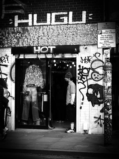 Black and white. A man stands in the open doorway of a Brick Lane fashion shop. He is very skinny and is wearing mid-calf length "shorts", well-above-the-ankle white socks, chunky black and white sports shoes, a white long-sleeved sweatshirt, a short-sleeved check shirt with a collar, and a dark beanie with a black star pattern. Painted above the doorway is the word, "HOT"...