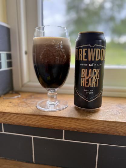 A black pint can with gold letters is next to a tall Stella Artois glass near a window