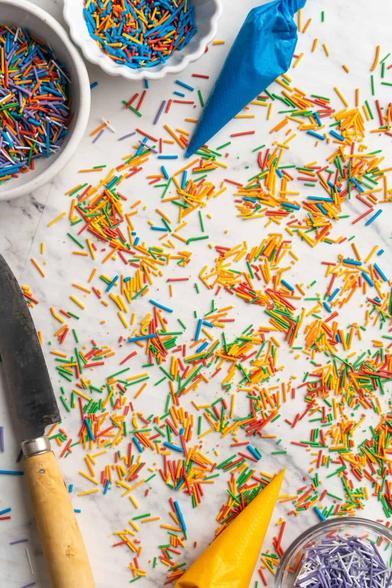 "Colorful rainbow sprinkles scattered across a marble surface, with bowls of sprinkles, a serrated knife with a wooden handle, and two piping bags, one blue and one yellow, around the edges.