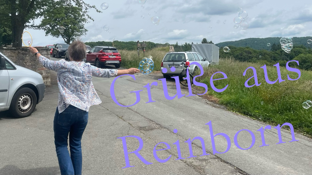 Eine Frau auf einem Weg hat die Arme gehoben; in einer Hand hält Sie einen Ring, an welchem sich Seifenblasen gebildet haben. Seifenblasen sind in der Luft. Text:“Grüße aus Reinborn“
