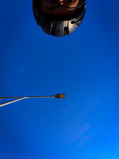 The vast majority of the image is dominated by a clear, vibrant blue sky with no clouds, creating a striking backdrop. At the left of the frame, a street light on a pole extends horizontally into the blue sky. A person wearing a dark helmet and glasses is looking down towards the viewer from the top of the frame.