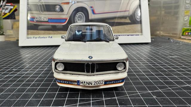 Model of a BMW 2002ti by Hasegawa in white in front of the framed box art, both on top of a cutting mat on a workbench.
