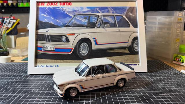 Model of a BMW 2002ti by Hasegawa in white in front of the framed box art, both on top of a cutting mat on a workbench.