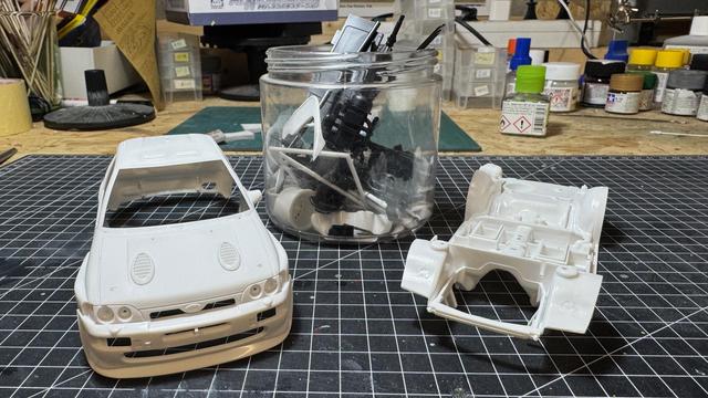 Parts of a Ford Escort RS Cosworth 4x4 by Tamiya on top of a cutting mat on top of a workbench. The body is on the left side, the chassis on the right, and a plastic cup with every other part in between.