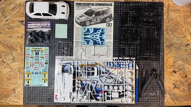 All parts of a Ford Escort RS Cosworth 4x4 by Tamiya layed out on top of a cutting mat on top of a workbench, including decals and the instruction manual.