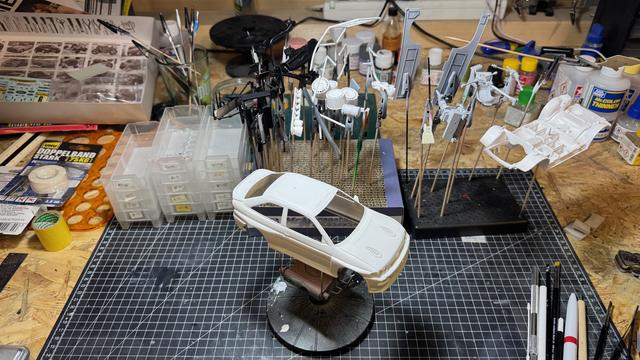 Parts of a Ford Escort RS Cosworth 4x4 by Tamiya on top of a cutting mat on top of a workbench. The body is on a painting stand, all other parts are clamped on painting clamps waiting for painting.