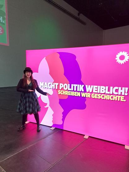 Liliane steht vor einer leuchtenden Aufstellwand mit dem Text "Macht Politik weiblich! Schreiben wir Geschichte."