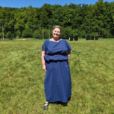 A white middle aged woman wearing a dark blue wool tunics, standing in a green field, with green woods behind.