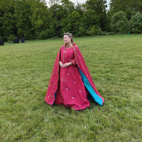 A white middle aged woman wearing a red silk gown with a red silk cloak lined in peacock blue silk. She has a crown on her head, and she is standing in a field with the woods behind her.