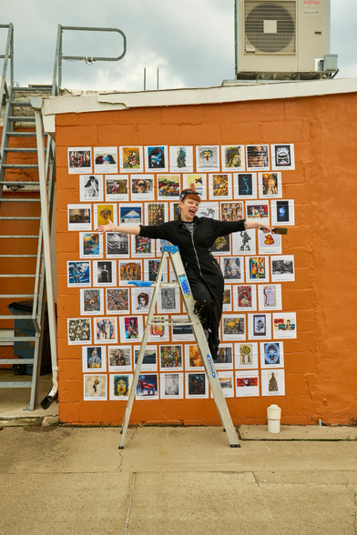 Perched atop a ladder, wearing a black speed suit, curator Klaire Lockheart hams it up by stretching their arms wide in front of large wheat-paste installation of epic prints on an orange building.