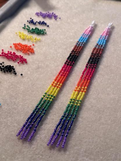 A pair of long beaded dangle earrings in the progress pride flag colors -- white, pink, light blue, brown, black, red, orange, yellow, green, dark blue, and purple. The earrings are laying on a bead mat next to piles of seed beads in the same colors.