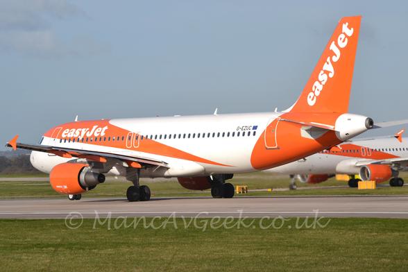 Side view of a twin engined jet airliner rolling from right to left along a grey runway.
The plane is mostly white, with a thick orange stripe running from over the top of the forward fuselage, down and back , to meet the lower rear fuselage just aft of the wing.
The front portion of this orange stripe has white "EasyJet" titles.
If anything, it looks like this plane is wearing a bandana.
The plane has a bright orange rear fuselage and tail, with white "EasyJet" titles, while the engine pods slung under the wings are also bright orange, as are the vertical wingtips.
Green grass fills the foreground, lining the side of the runway, with more grass in the background running off into the distance.
Another jet airliner in a similar livery is behind this planes tail on the right of the frame.
Grey-blue sky fills the rest of the frame.