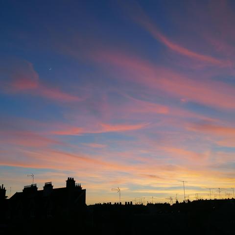 Photo carrée d'un ciel au soleil couchant. Le ciel est bleu, strié de nuages qi vont du jaune au rouge, de bas en haut. Sur un cinquième de la photo en bas, on voit des toits, cheminées et antennes, tout est noir. Les couleurs du ciel et des nuages ressortent beaucoup. On aperçoit un fin croissant de lune en haut à gauche.