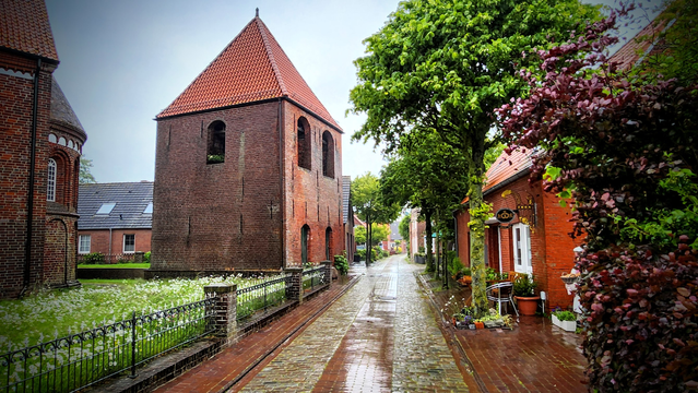 Ein nicht sehr hoher aber dafür recht breiter Glockenturm steht an einer kleinen Dorfstraße.