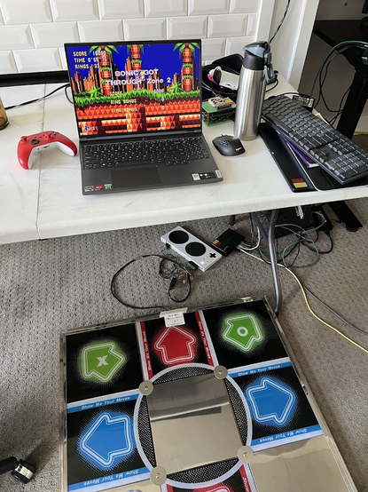 A metal-framed Dance Dance Revolution pad (colorful arrow buttons that a player presses with their feet, usually in sync with a rhythm game), connected to a 15-pin (VGA) breakout port, then hooked up to the inputs on an Xbox Adaptive Controller. That controller is then operating as an input to play Sonic CD on a laptop, currently showing the "Sonic got through Zone 2" end screen.