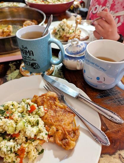 Tisch mit Geschirr, Besteck, Tassen, Zuckerdose, Schalen mit Hühnchen und Coucous-Salat. Rechts sieht eins eine Hand, die eine Gabel hält, der Teller ist gefüllt. Die Person trägt ein rosa Pokémon Tshirt. Im Vordergrund ein weißer Teller, auf ihm liegt rechts Besteck, in der Mitte liegt Couscoussalat und braun gebackenes Hühnchenfleisch.