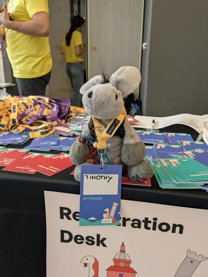 Plush donkey is sitting on the conference registration desk and proudly showing off his shiny new conference badge saying "Timothy" on it.