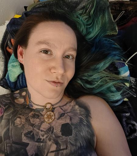 pyrrh laying down on colorful blankets with a little smirk and sparkles in its eyes. its hair is long and wavy in a greenish color, and its chest tattoo of a fragmented wolf is prominently visible