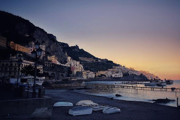 A picturesque view of the Amalfi Coast at golden hour, with the sun setting behind the coastal cliffs. Buildings with colourful facades are built into the side of a steep hill, overlooking a calm sea. A pebbled beach in the foreground features several small overturned boats, while a pier with boats moored alongside extends into the water on the right. The sky transitions from a deep blue at the top to a warm orange and yellow near the horizon.
