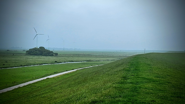 Von einer Deichkrone blickt man landeinwärts wo graue Wolken tief hängen und der Wind Sprühregen über das Land treibt.