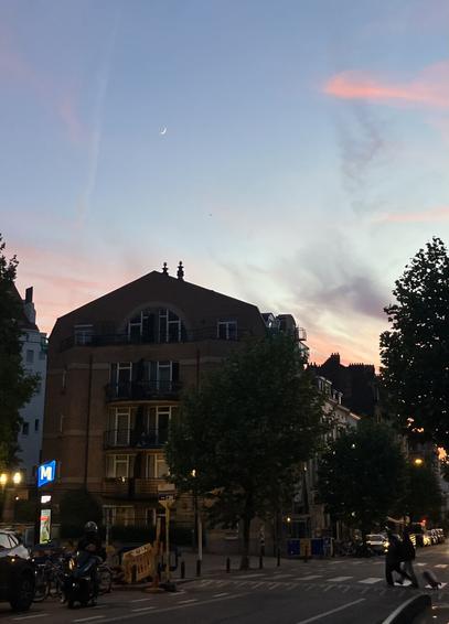 At the foreground on the right a metro station and a apartment block, not very high, behind, in the dark. On the right the same street, the two people on the zebra crossing, just their shapes are visible. The upper half of the picture shows the sky. The street lights are on but the sky is still quite clear with some pink clouds. The moon appears also, very tiny, just above the apartment block.