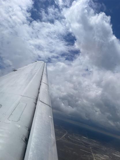 The wing of a plane stretching out to a cloudy sky over fields on a plain.