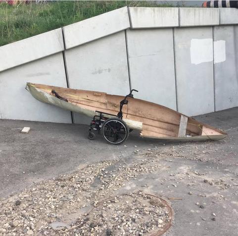 A dilapidated wooden canoe rests atop a wheelchair on a sidewalk.