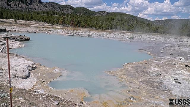 USGS research web camera image from Biscuit Basin in Yellowstone National Park today. The image shows Black Diamond Pool in the foreground, with a background of green trees and the slopes surrounding the basin. Rocks ejected in a 2024 hydrothermal explosion can be seen around the pool. This is a live view of an area of the park that has been temporarily closed to the public due to the potential for future hazardous events.